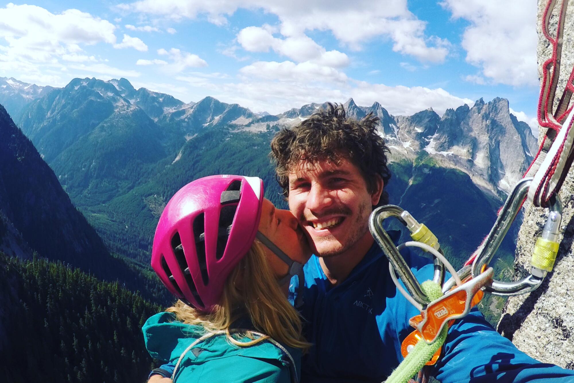 A woman wearing a pink helmet kisses a man as theyre both roped to the side of a mountain, with mountains in the background