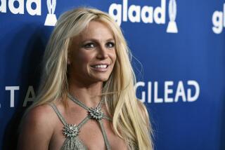 Britney Spears in a sparkling, silver strapped gown smiling and posing against a dark blue background