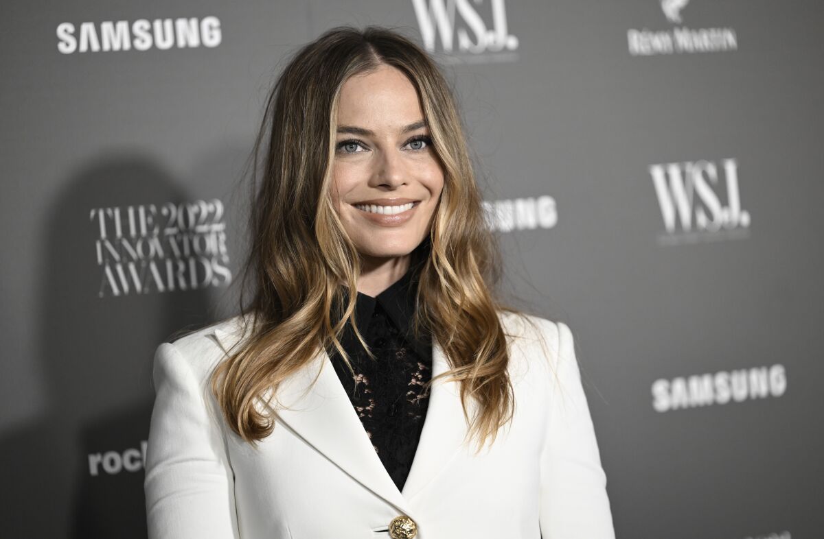A woman with long hair wearing a white blazer over a dark shirt smiles upon arrival at an event