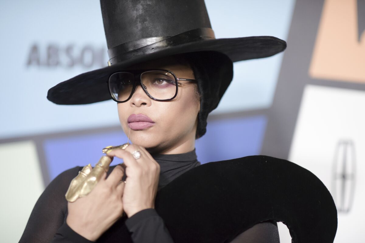 Erykah Badu, wearing a large black hat and rimmed glasses, clasps her hands in front of her.