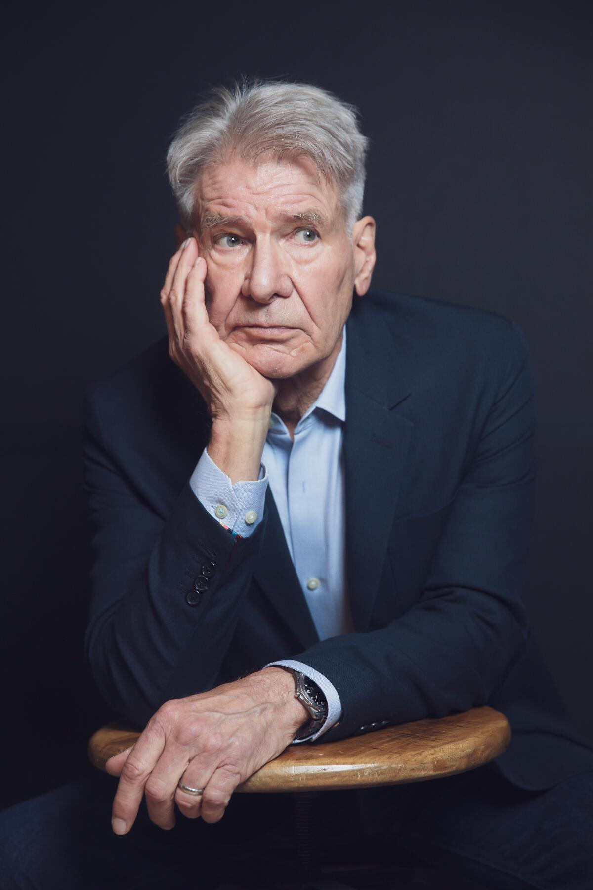 Harrison Ford holds one hand to his face and leans against a stool.