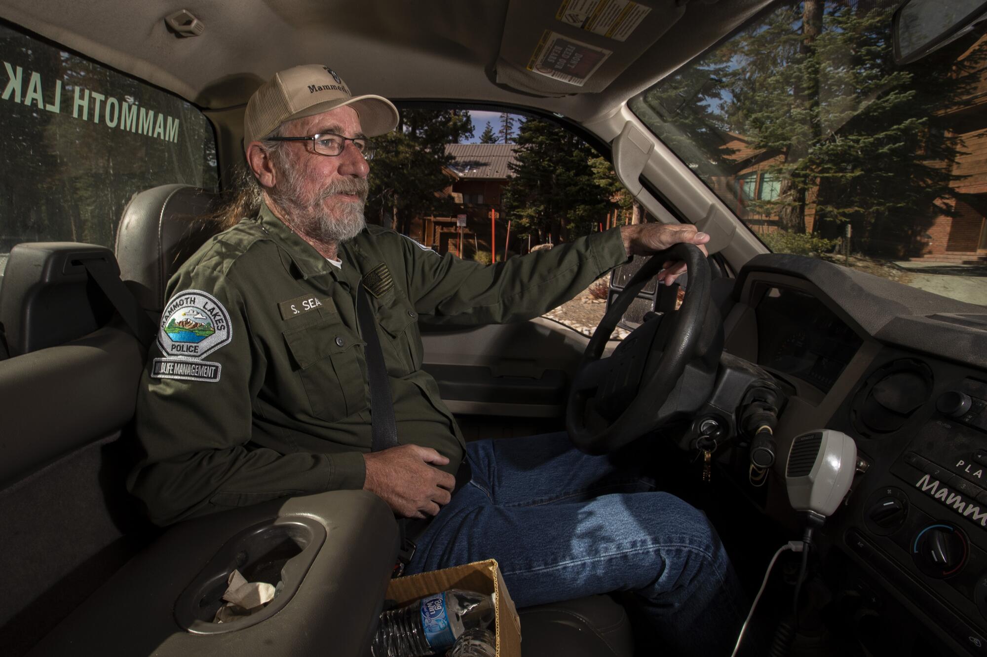 A bearded man in a cap and ranger jacket sitting in a truck.