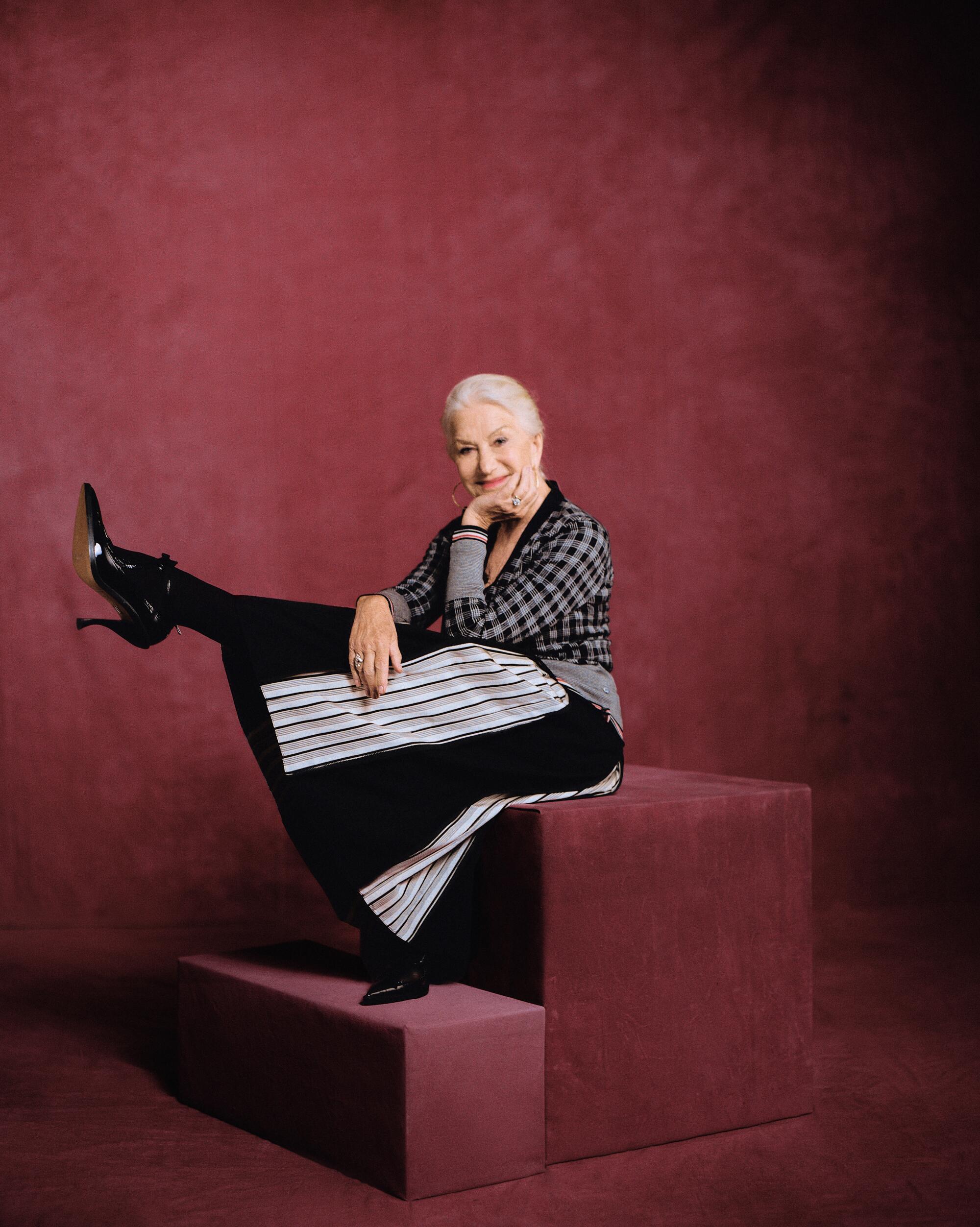 Actress Helen Mirren sits on a prop cube and kicks up her leg for a portrait.