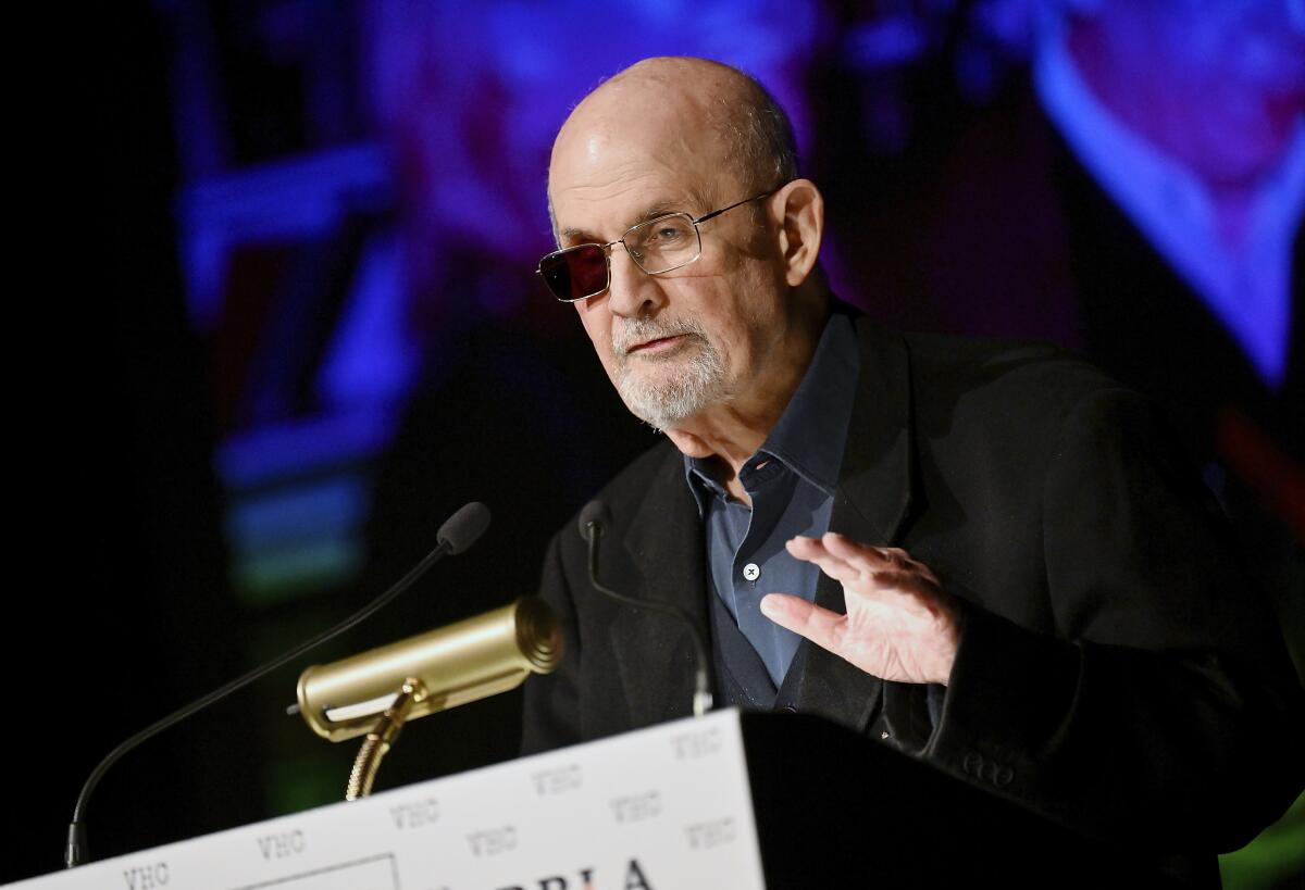 Salman Rushdie wearing glasses with one dark lens and speaking at a lectern
