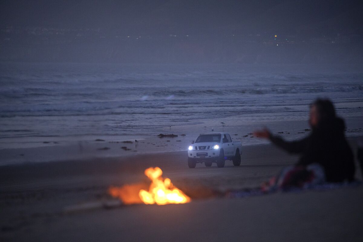 A truck drives on the beach with its headlights on as a person sits at a small bonfire in the foreground
