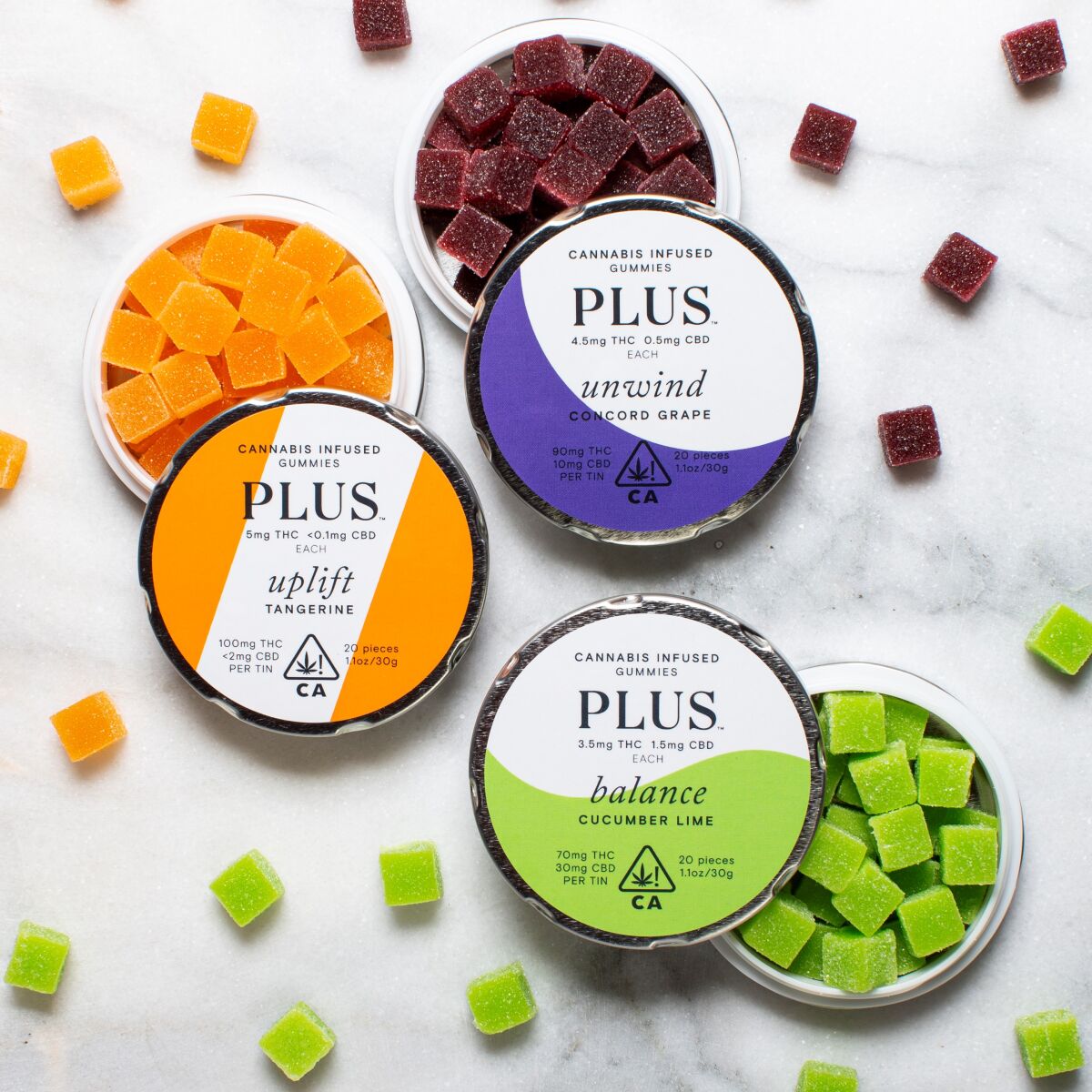 Three round tins containing Plus Products' small square cubes of cannabis edibles.