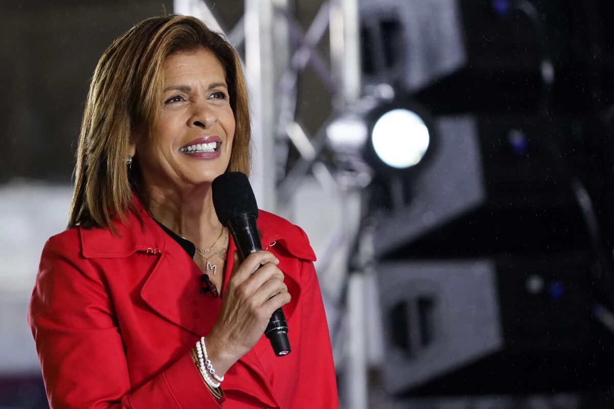 A woman with brown hair in a red suit holds a microphone and smiles under stage lights