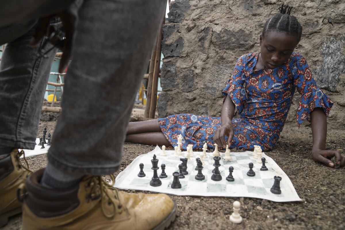 A girl sits on the ground and looks over pieces on a fabric chess grid. 