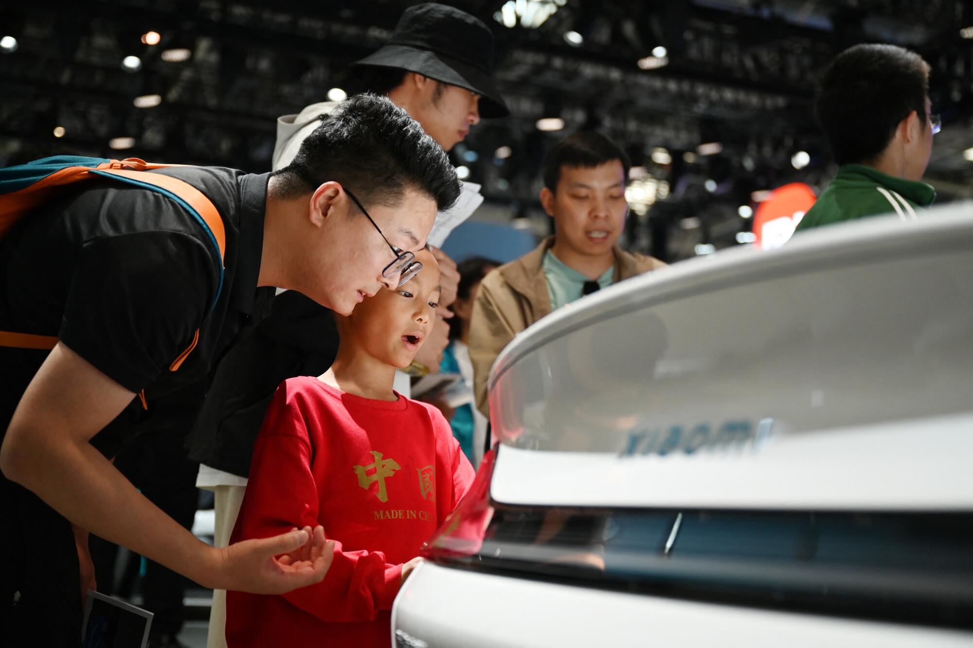 A person in a dark shirt and glasses leans next to a boy in a red t-shirt looking at a vehicle. 