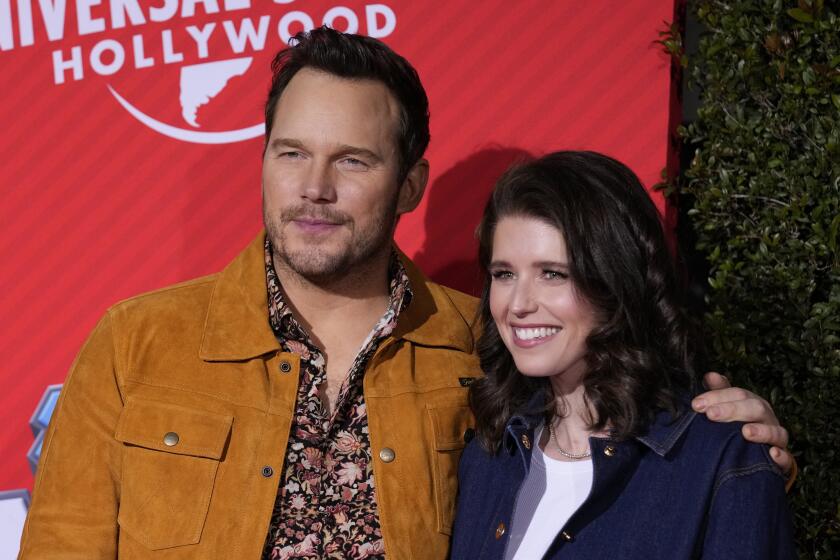 Chris Pratt in a brown jacket and a floral pattern shirt posing next to a woman with dark hair in a dark jacket