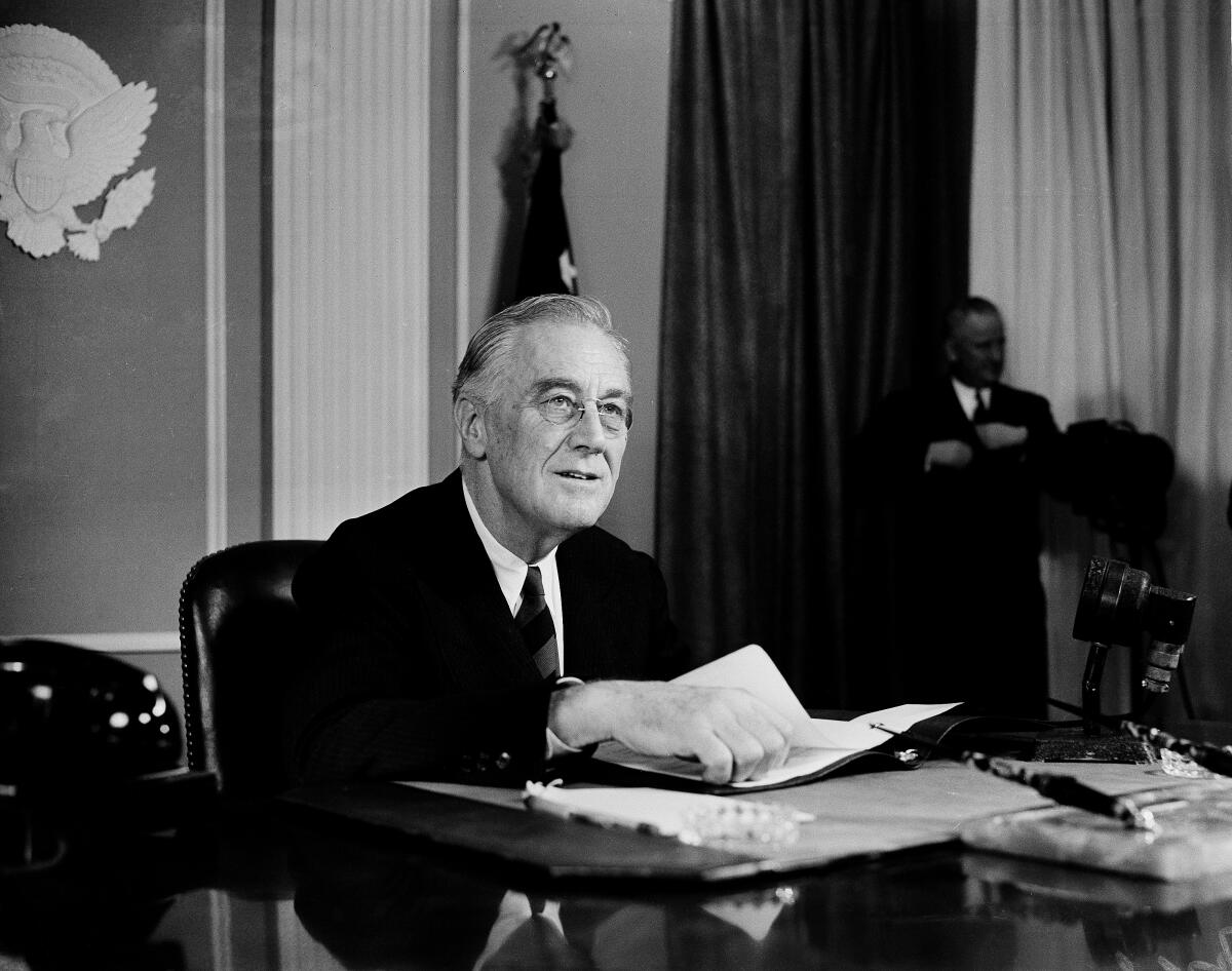 Roosevelt, in suit and tie, sits at a desk with one hand on some papers.