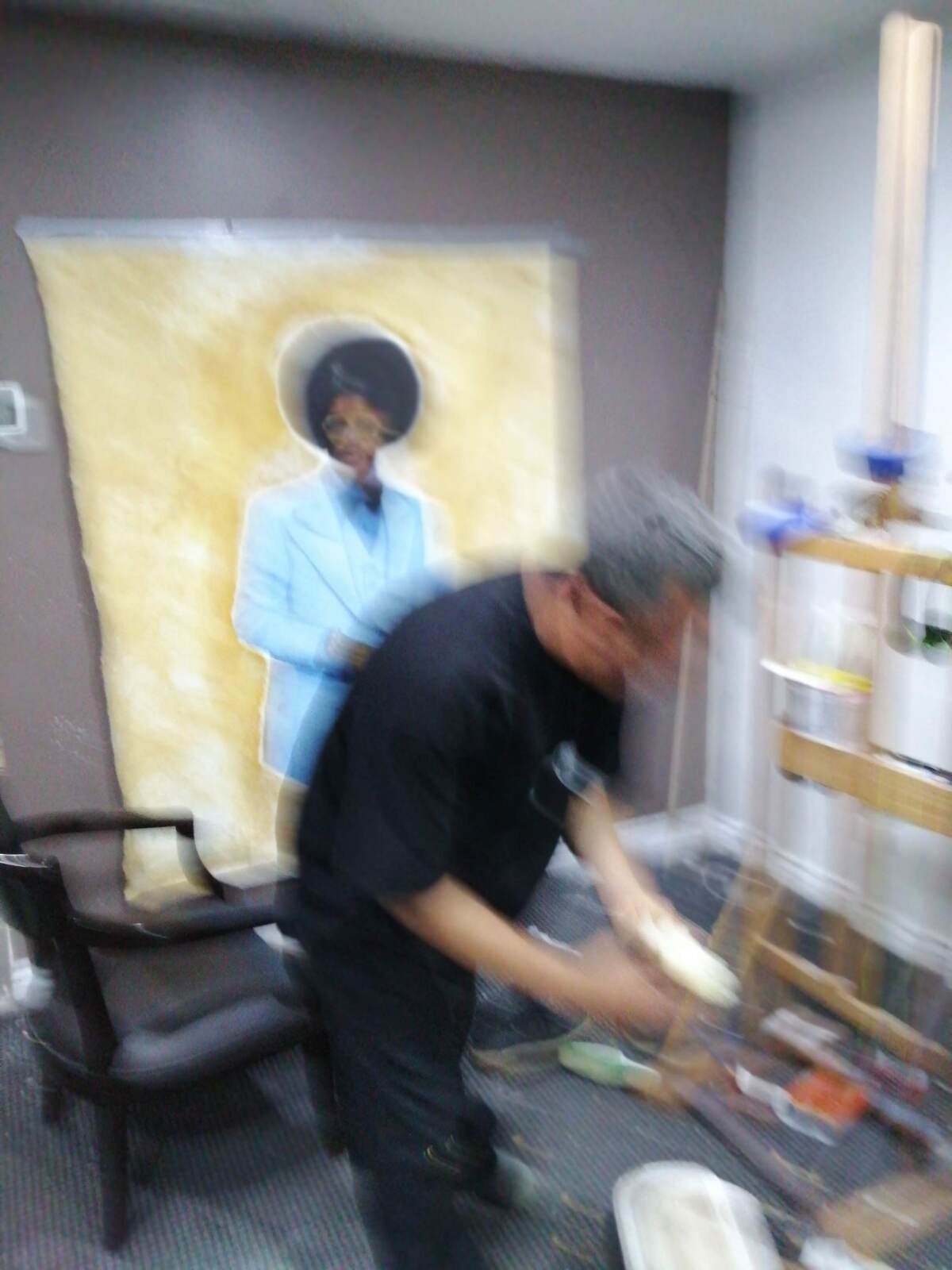 A blurry view of a man with his back to a painting in progress of a Black man in a blue suit against a mustard background 