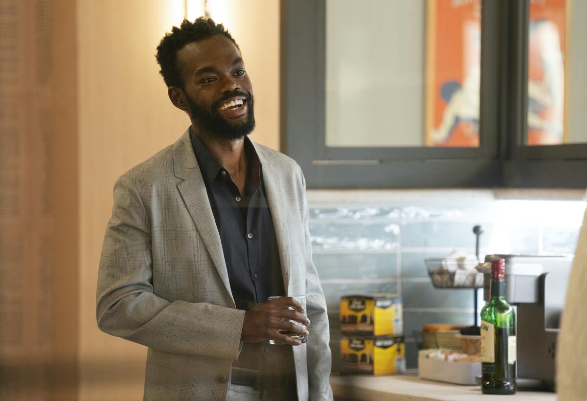 A man in a gray blazer stands in a kitchen holding a drink.