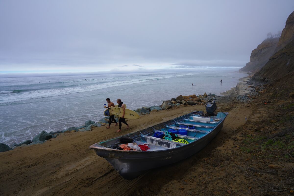 Two people carrying surfboards walk past a panga boat on a beach on a foggy day