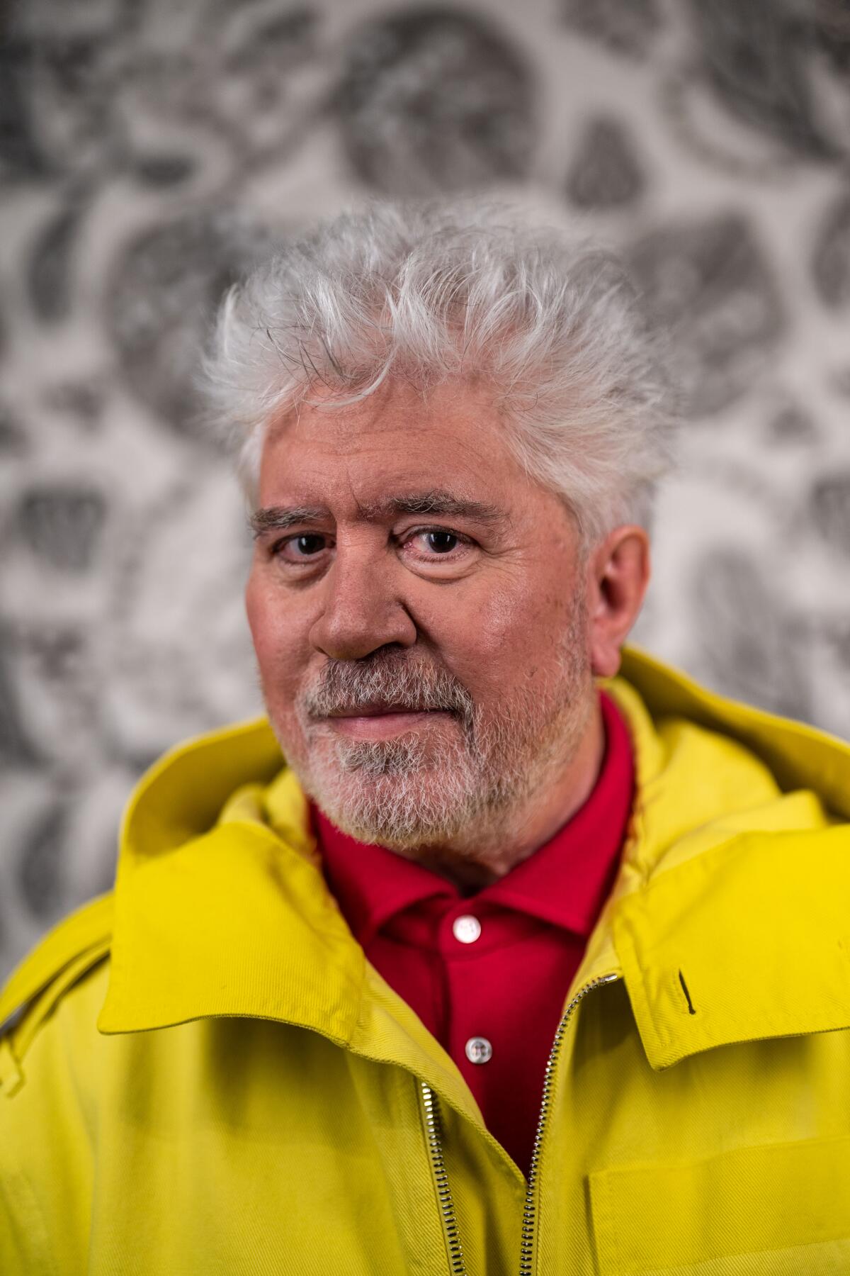 Pedro Almodóvar, in a red collared shirt and bright yellow jacket, looks into the camera.