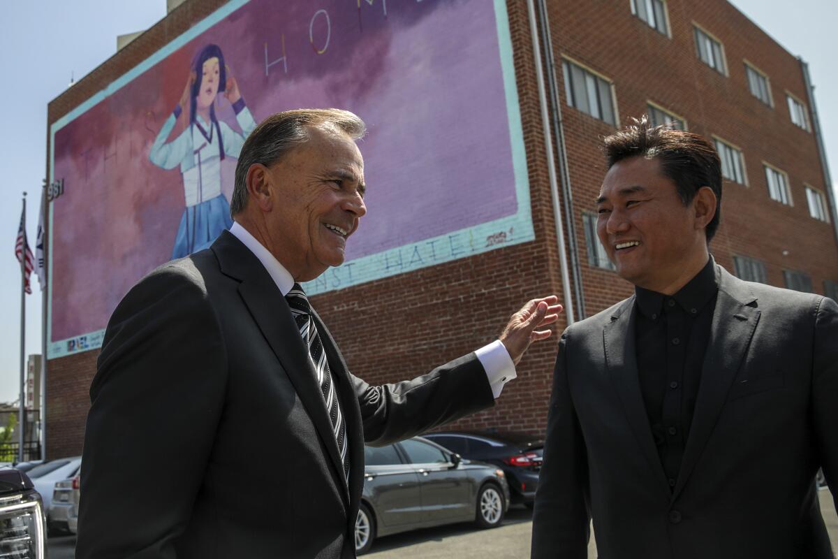 Two men in suits smile outside a building with a large mural
