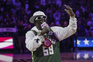 Flavor Flav in a white sideways hat, sunglasses, jacket and jersey singing into a microphone with his left arm up