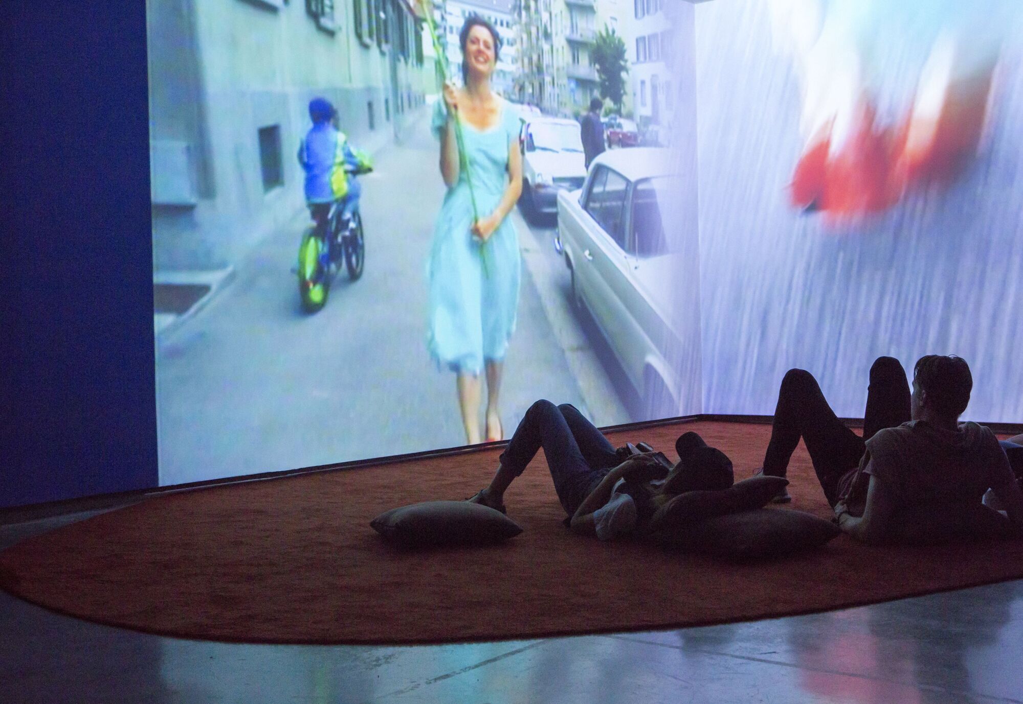 People lay on pillows and a carpet to watch a wall-sized video piece occupying an entire gallery corner