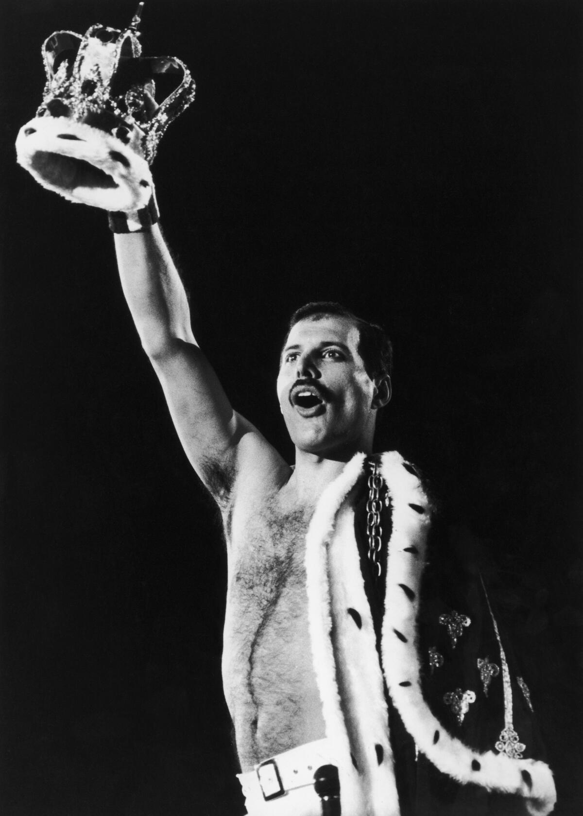 Freddie Mercury holds a crown up to the sky, with a cloak draped on other side of body