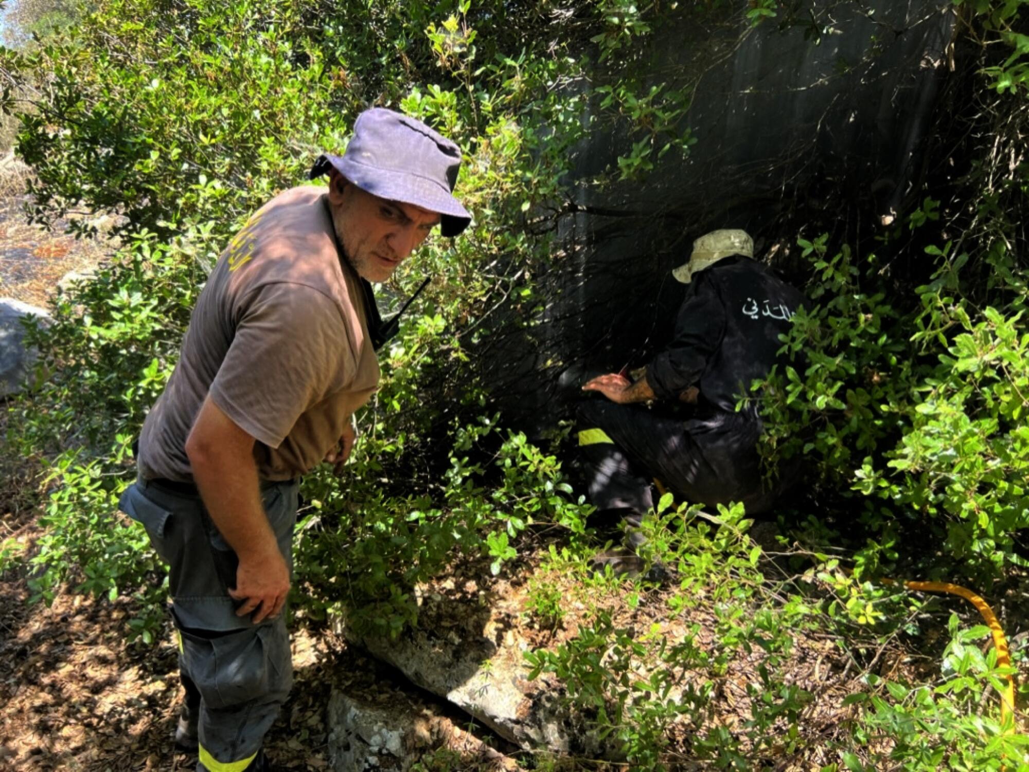 Men wearing floppy hats put out a small fire in a brushy, wooded area.