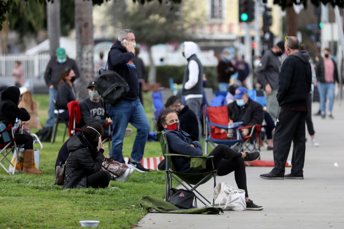 People wearing masks sit in folding chairs on the sidewalk and on the grass.