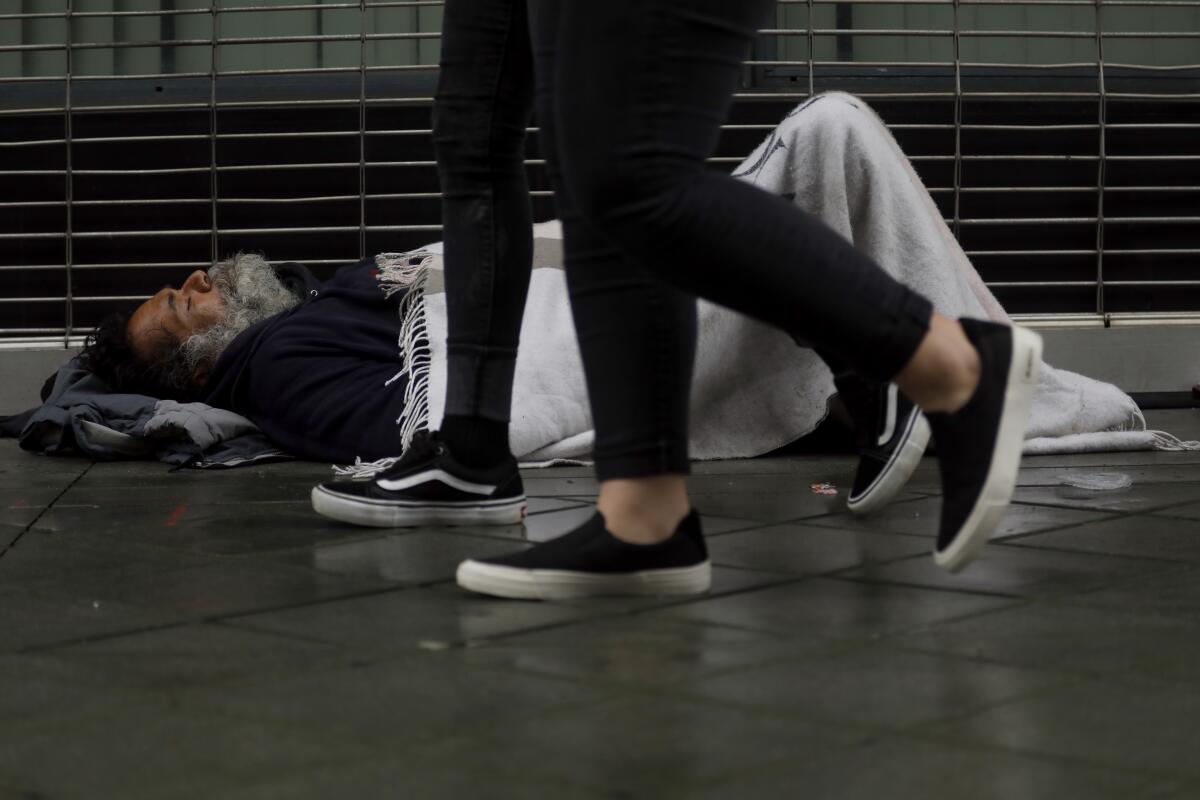 Cubierto con una manta, una persona sin hogar trata de mantenerse caliente mientras los peatones pasan junto a él en el centro de Los Ángeles.