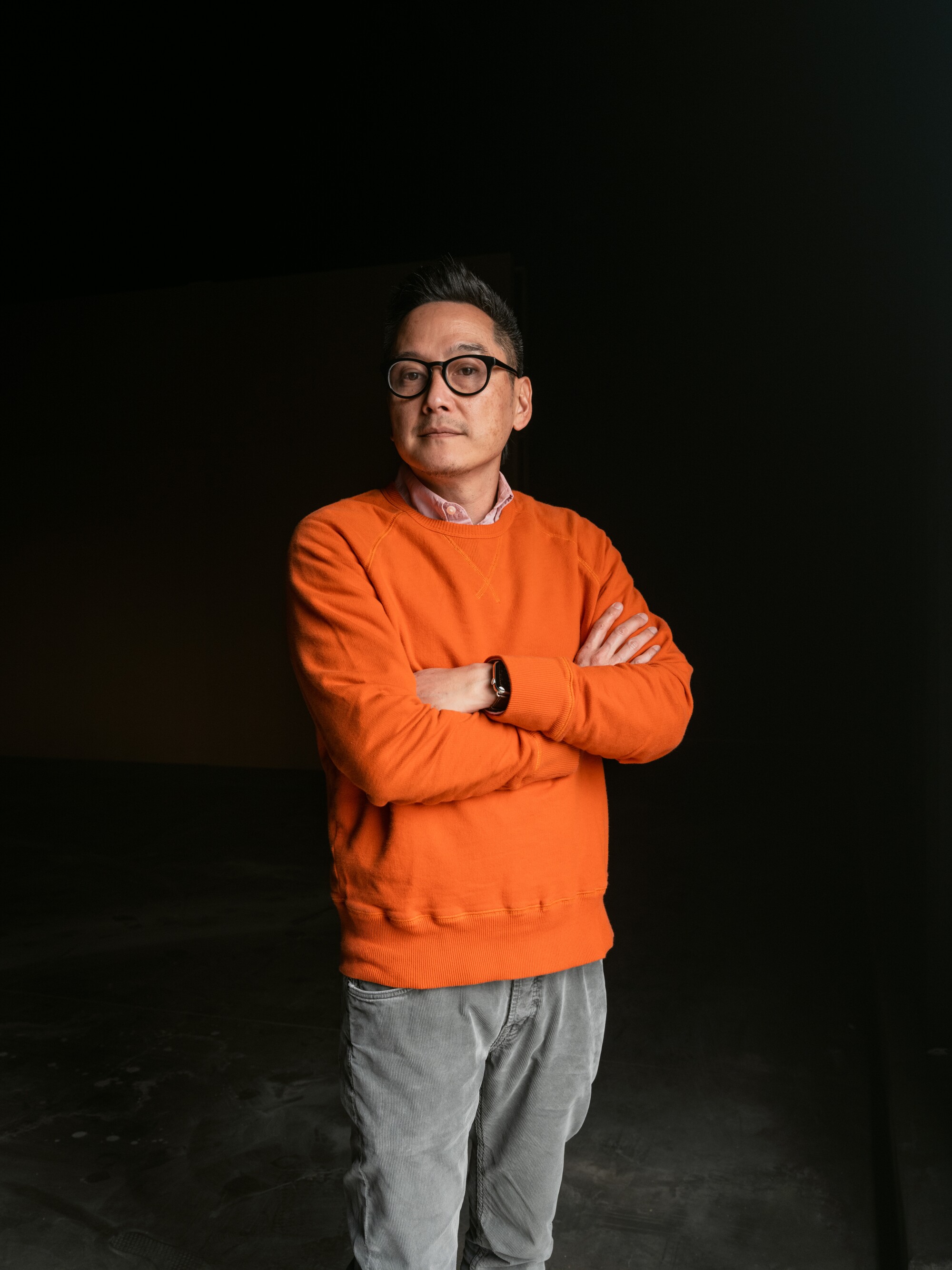 A man in an orange sweater stands with his arms crossed 