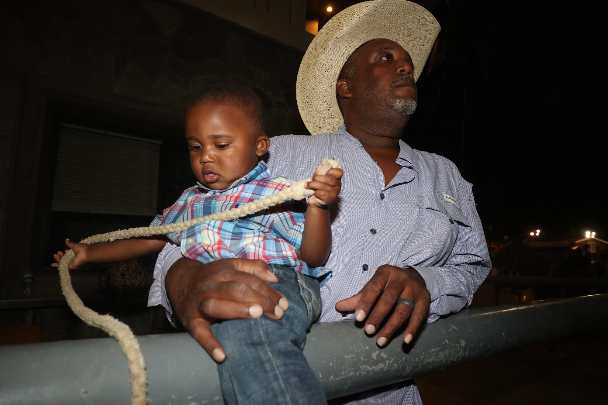 A young boy plays with a rope as he is being held by a man in a cowboy hat.