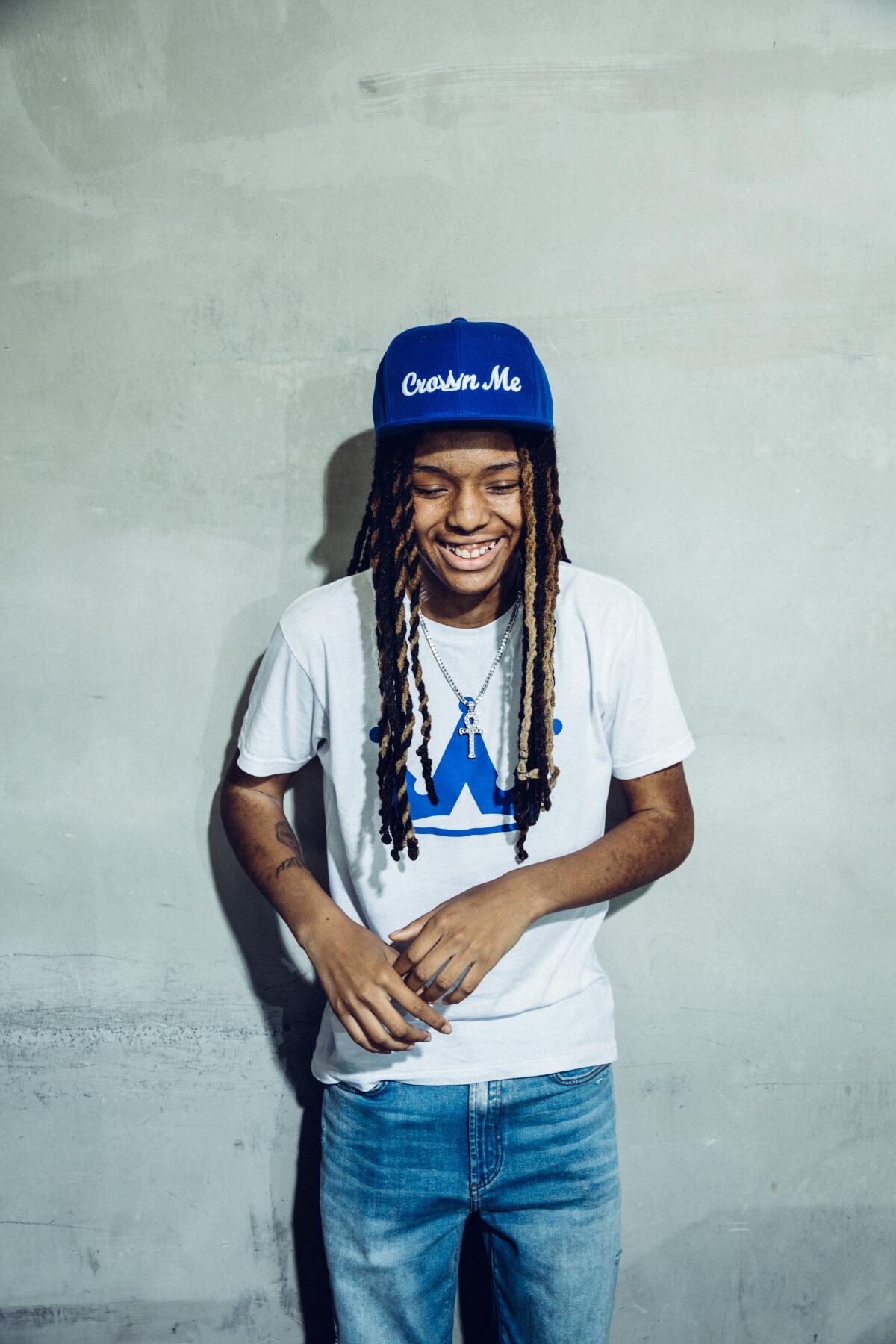 A teen actor in a baseball cap that reads "Crown Me" poses in front of a white wall.