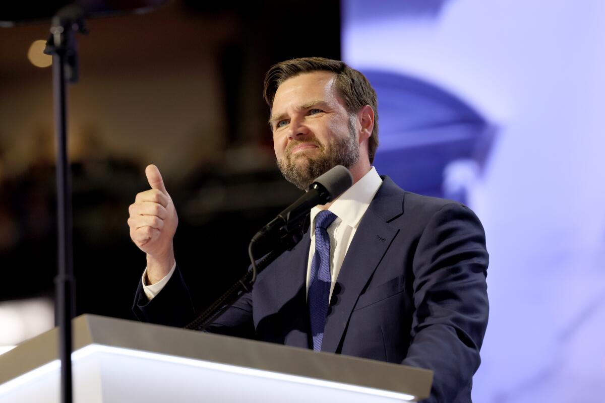 A white man with brown hair and a brown beard gives a thumbs up gesture from behind a lectern