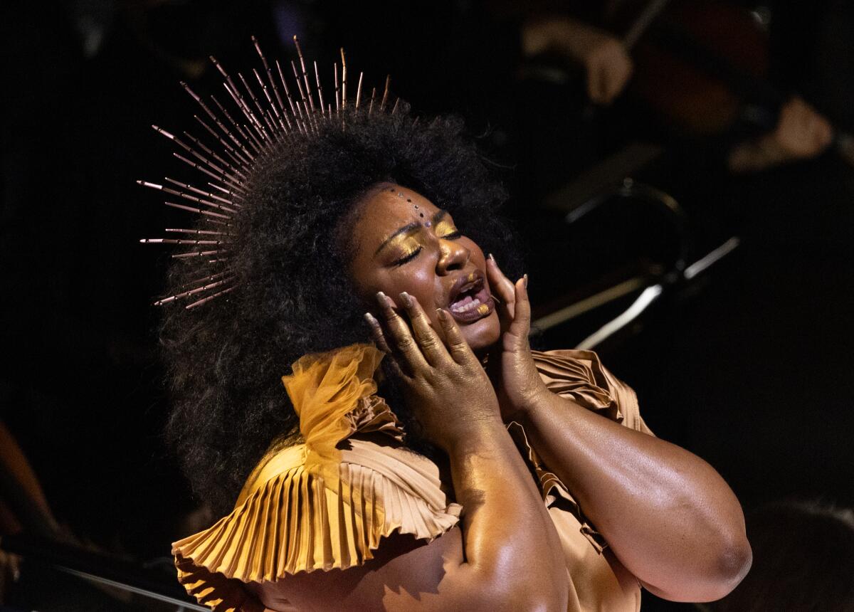 Raehann Bryce Davis, a Black woman in a gold dress with a gold crown, closes her eyes and rests her hands on her cheeks.