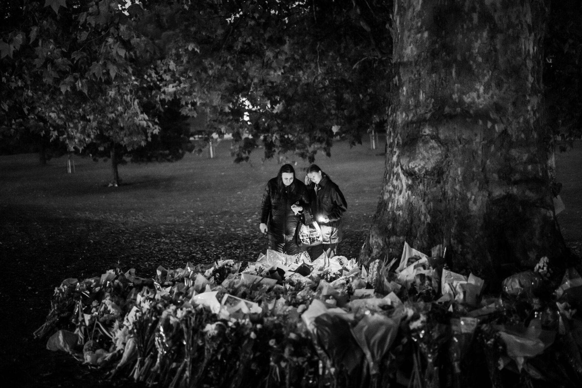 Two people stand with a candle by a pile of flower bouquets under a tree.