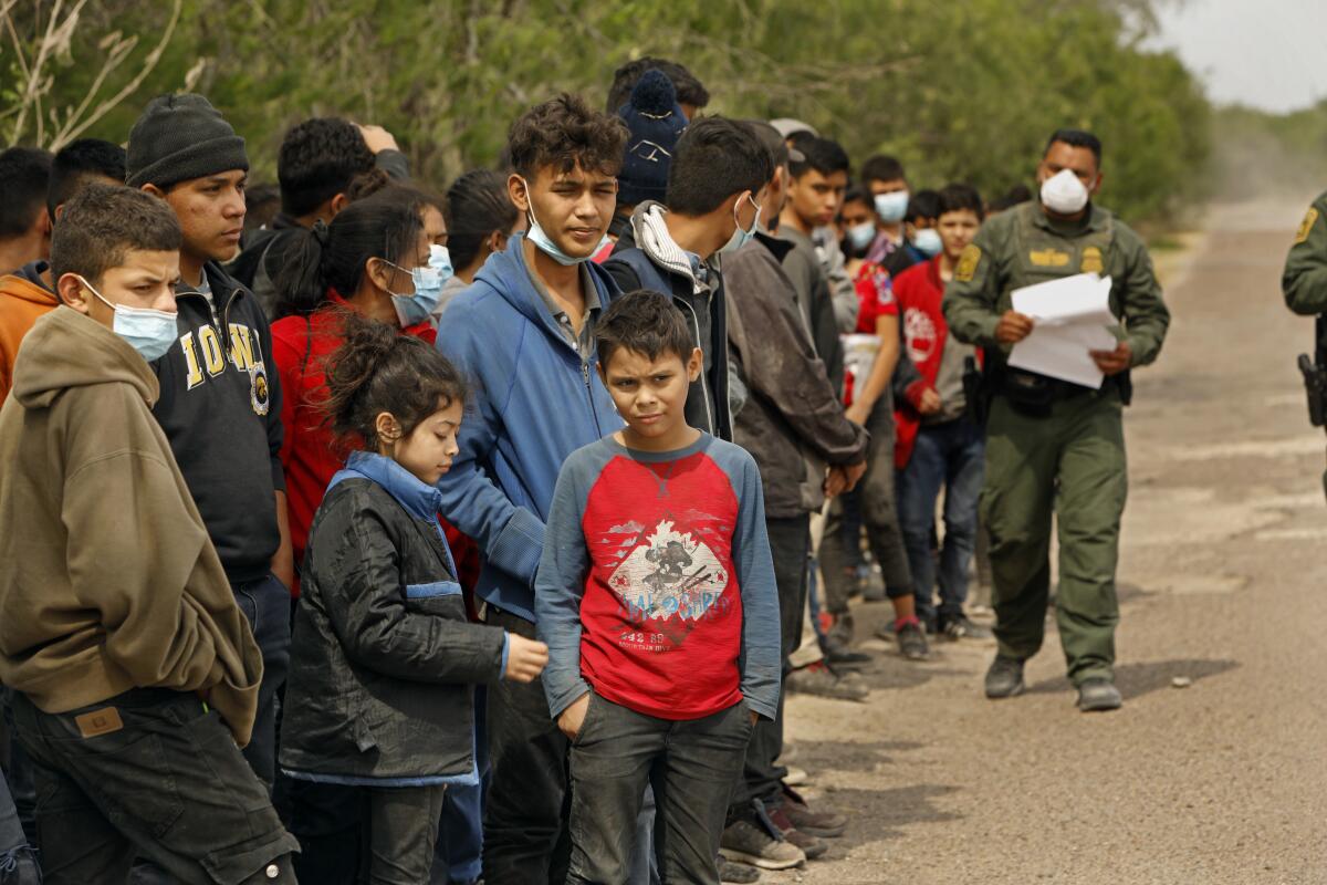 Young people stand outdoors in a line alongside a masked man in a uniform holding papers. 