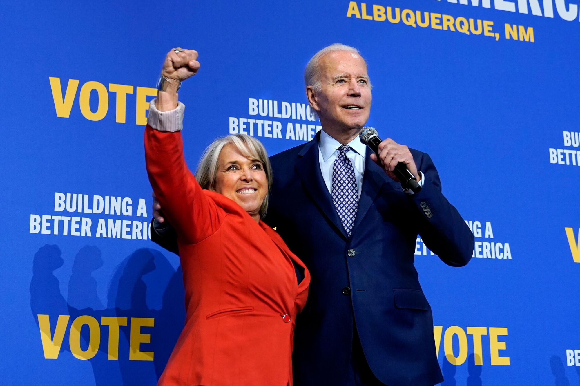 A woman with blond hair, in a red suit, raises a fist next to an older man in dark suit and striped tie holding a microphone 