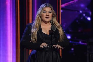 Kelly Clarkson with dark makeup in a black long-sleeve dress standing on a stage speaking into a microphone