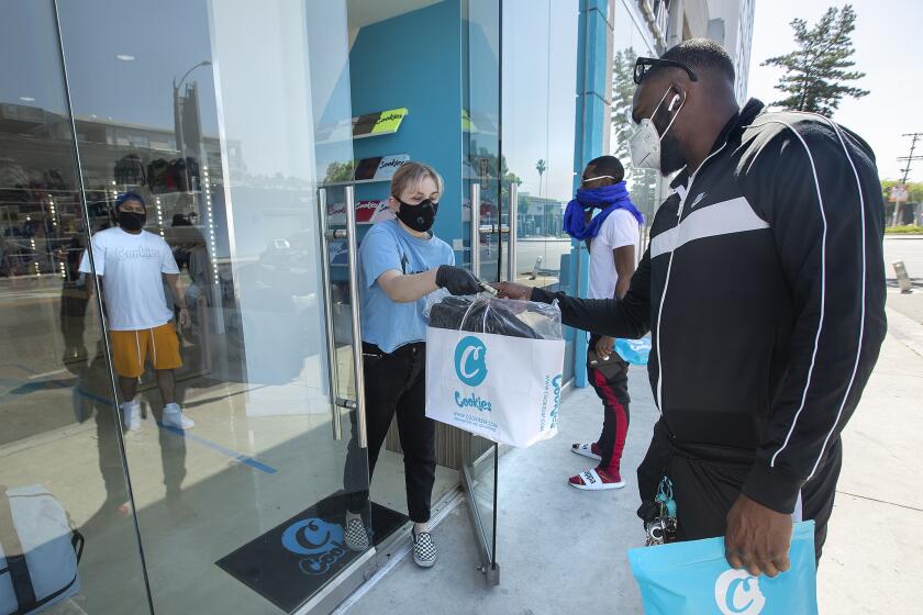 LOS ANGELES, CA-MAY 8, 2020: Jessica Knudsen, center, a sales floor associate at Cookies, a street wear clothing store on Melrose Ave. in Los Angeles, hands over change and a bag containing a backpack purchased by customer Jason Green, of Tampa, Florida. In background, left, is store manager Angel Lopez and 2nd from right is customer Jay Pooka of Atlanta, Georgia. Today was the first day the store re-opened since being forced to close due to the coronavirus outbreak. The store offered curbside shopping only to customers. (Mel Melcon/Los Angeles Times)