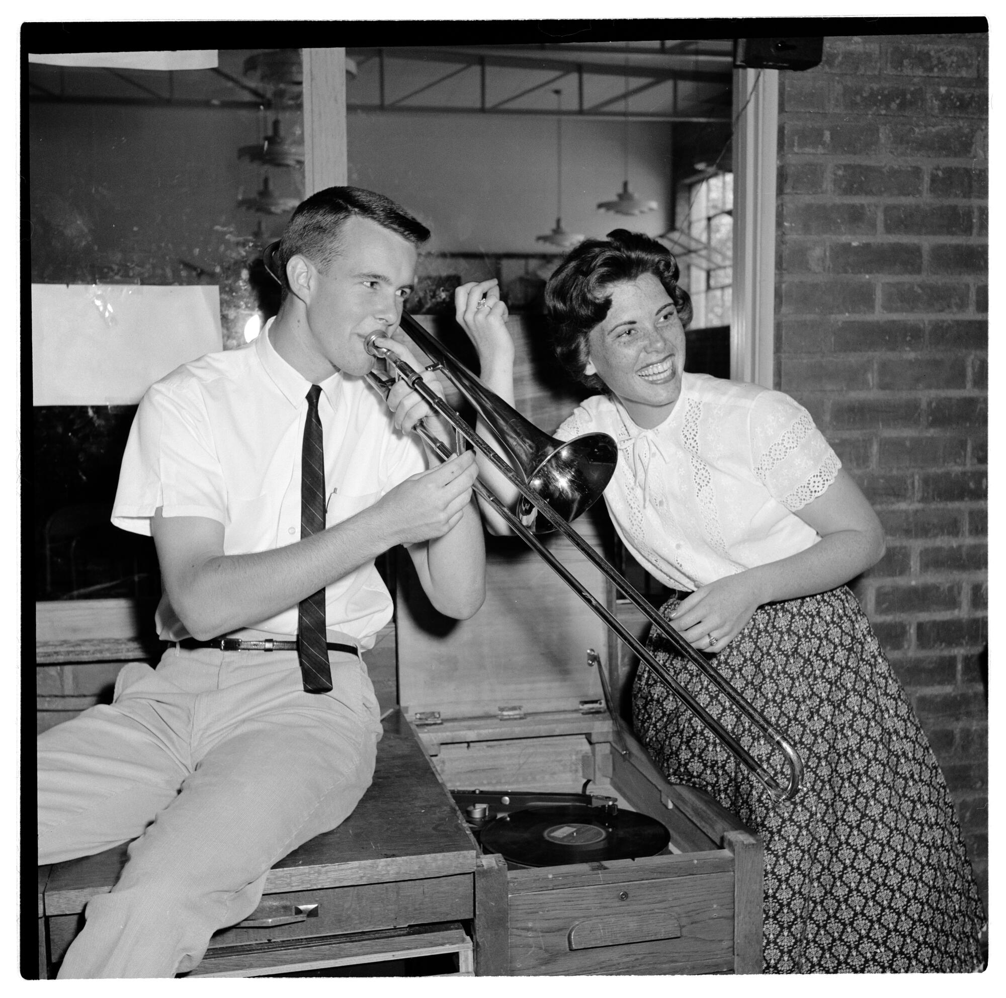 A young white man plays the trombone while a young white woman sitting next to him snaps her fingers.