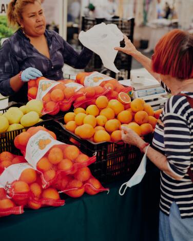 A red-haired woman buys citrus fruit from a woman at a farmers market stand.