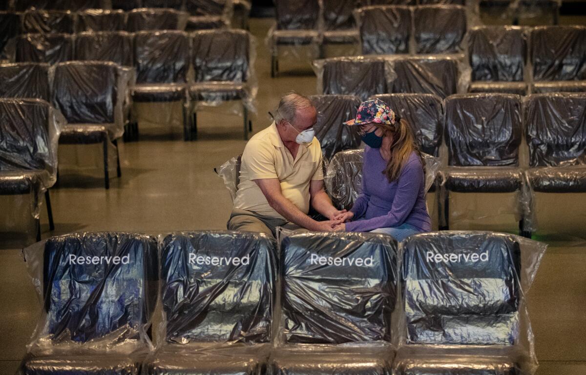 A man and woman hold hands and pray among plastic-covered chairs in a church.