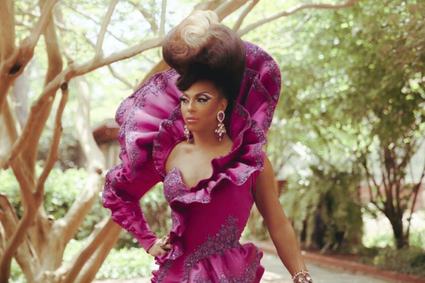 A drag queen in a high collar magenta gown standing in a grove of trees