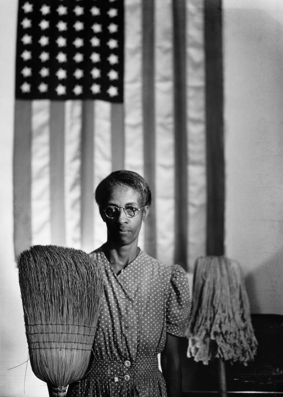 Ella Watson looks into the camera while holding a broom, an American flag behind her, in Gordon Parks' "American Gothic"