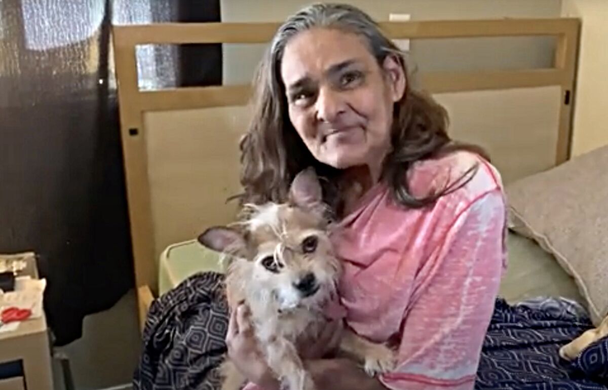 A woman sits on a bed while holding her dog in her arms