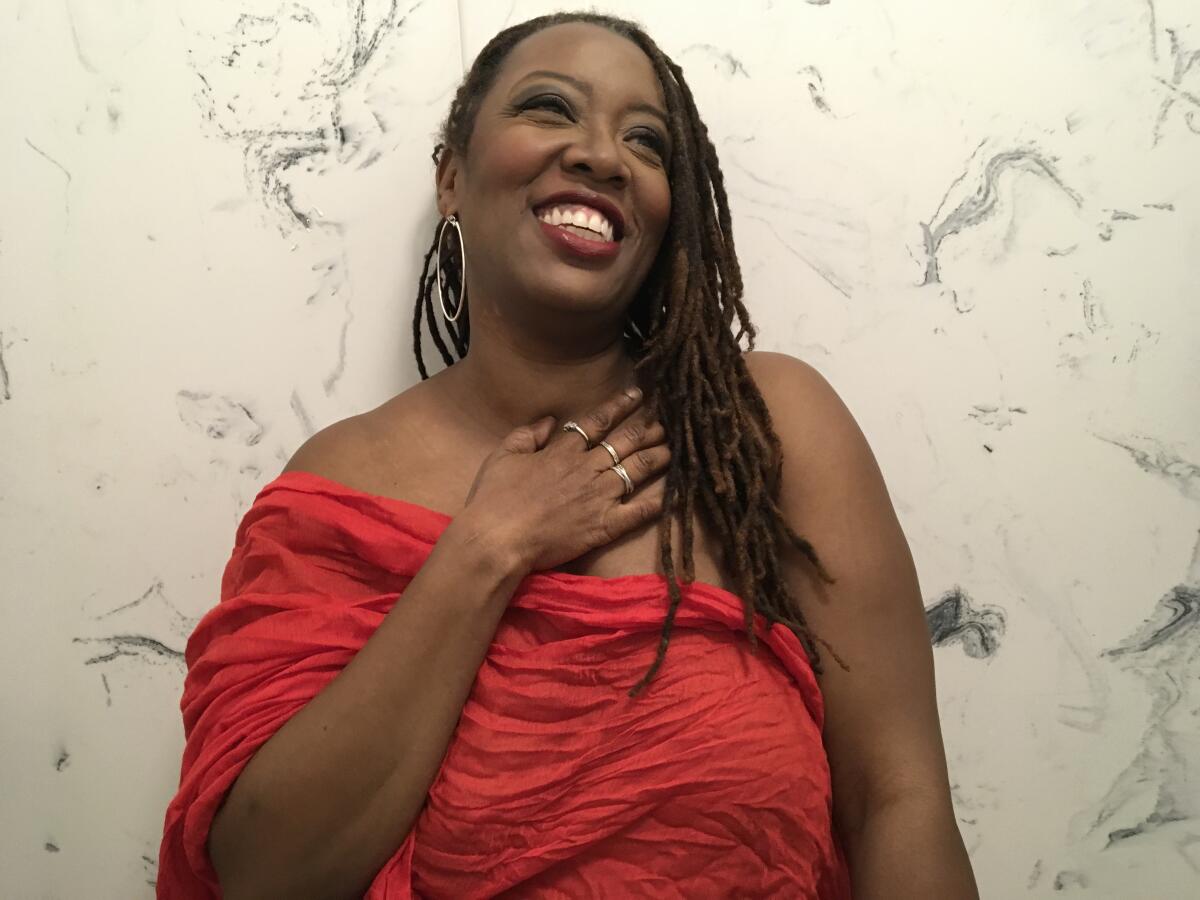 A woman with braided hair and hoop earrings smiles while holding her hand to her chest. 