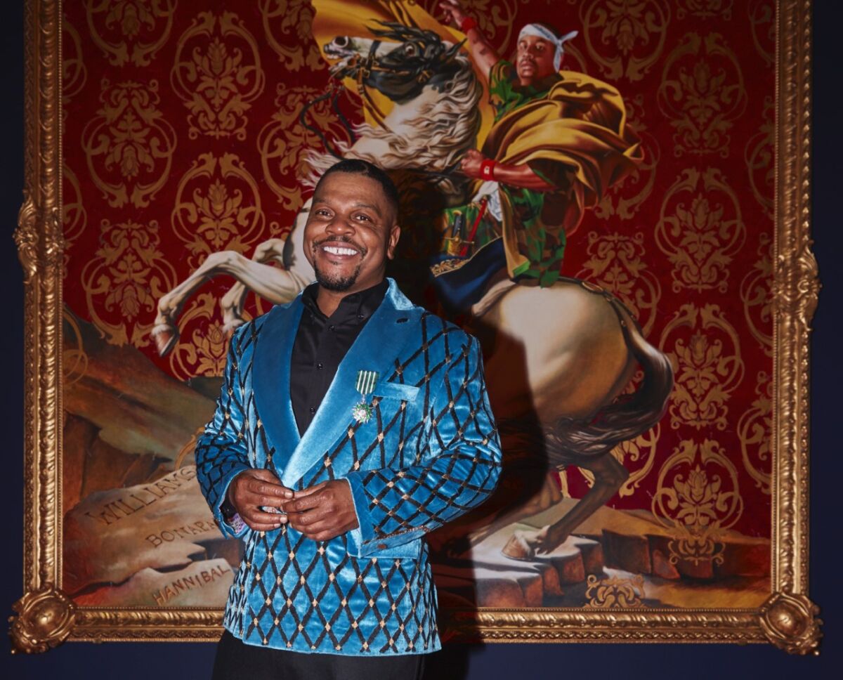 A man in a blue satin jacket stands in front of a large painting of a man on a horse