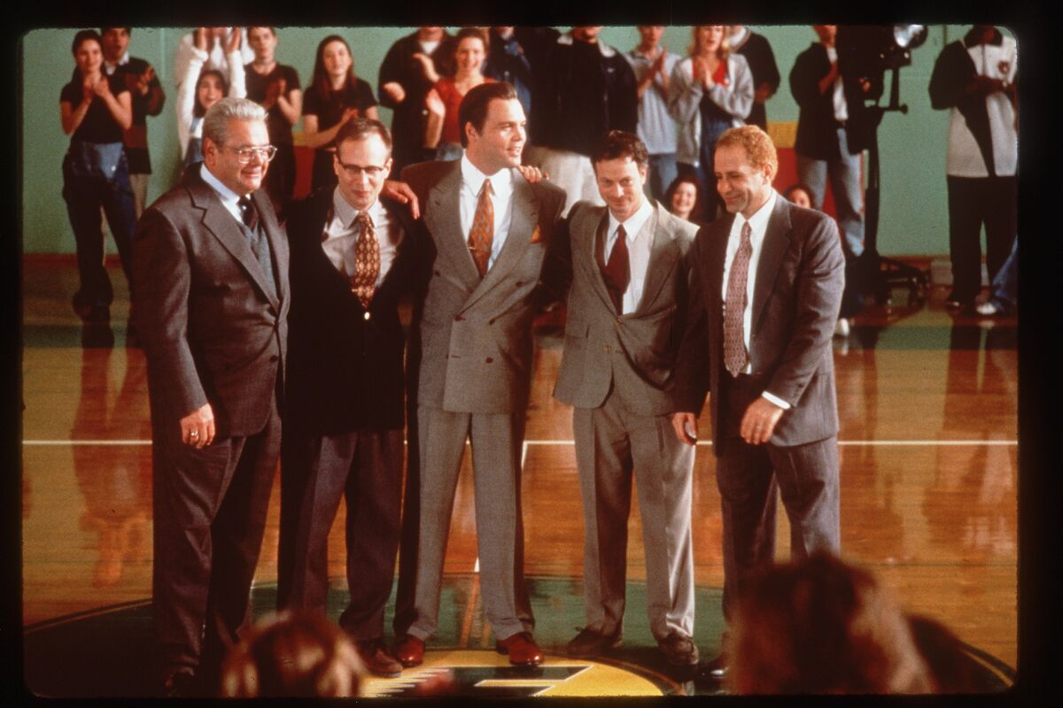 Five men in suits pose together in a gymnasium in a scene from a movie