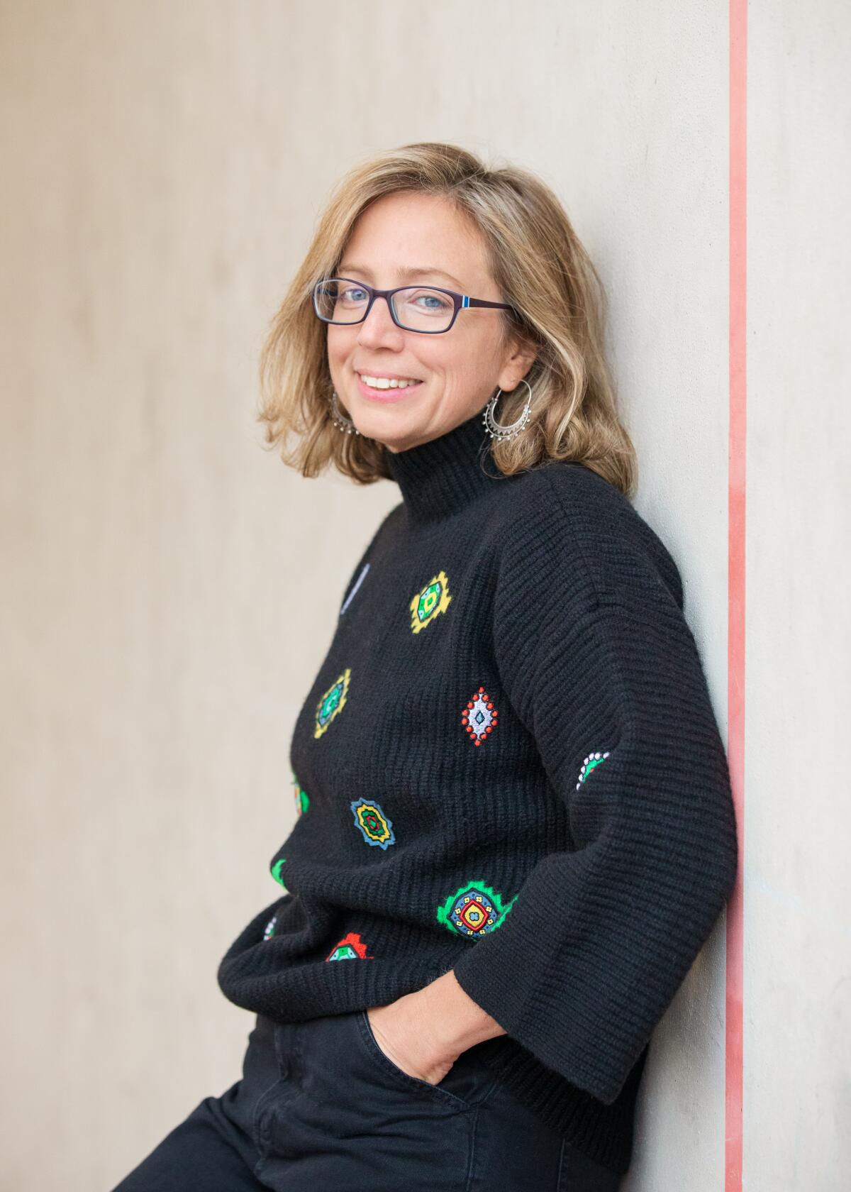 Alexandra Lange, wearing a black patterned sweater, smiles as she leans against a wall.