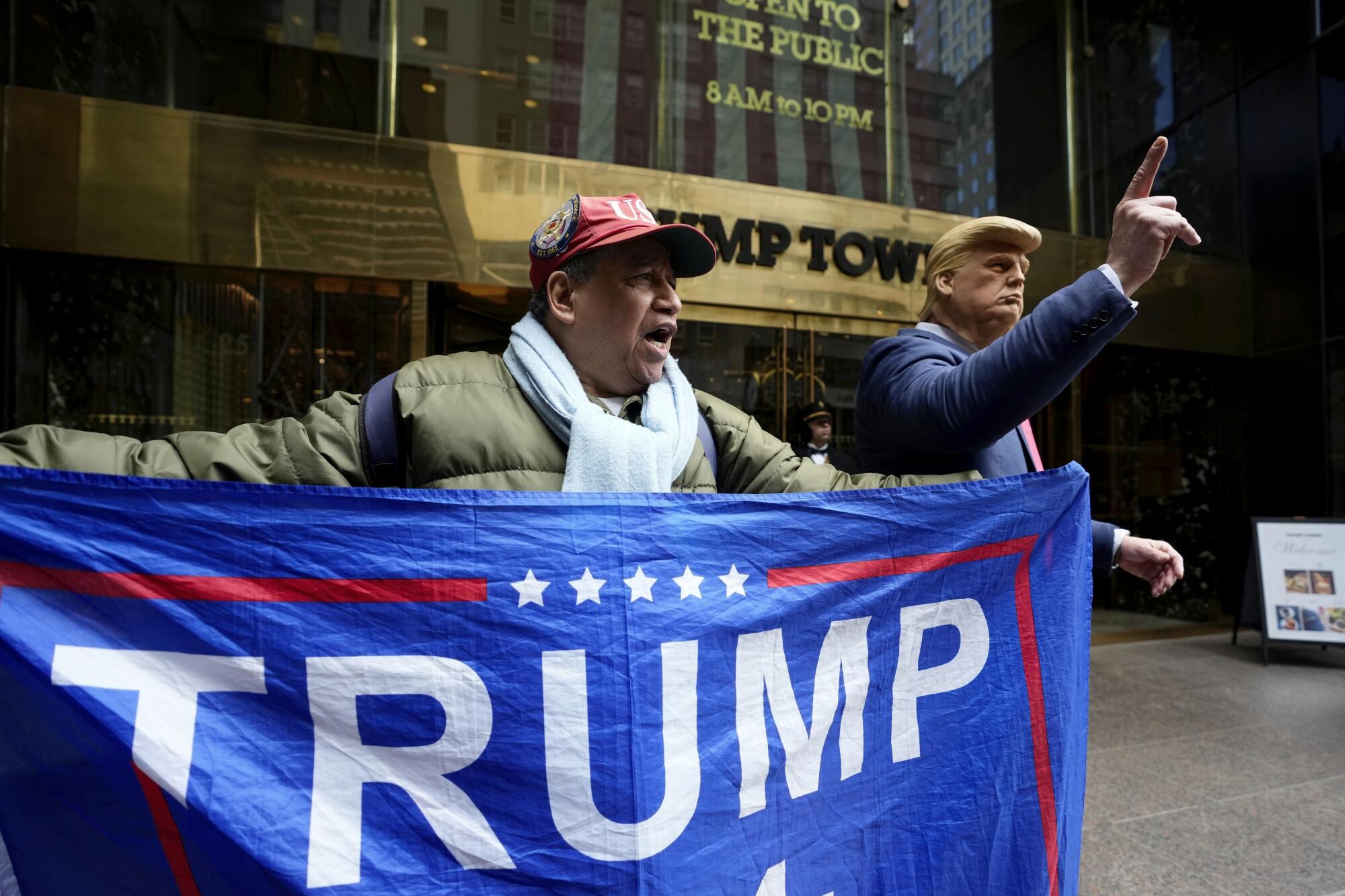 A man holds a Trump flag next to someone dressed as former President Donald Trump.