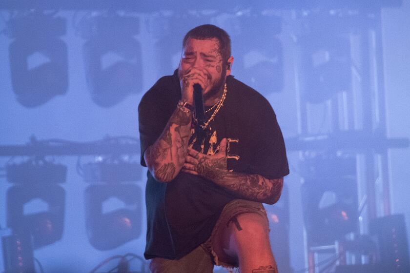 A man with tattoos on his arms and face crouches while singing onstage