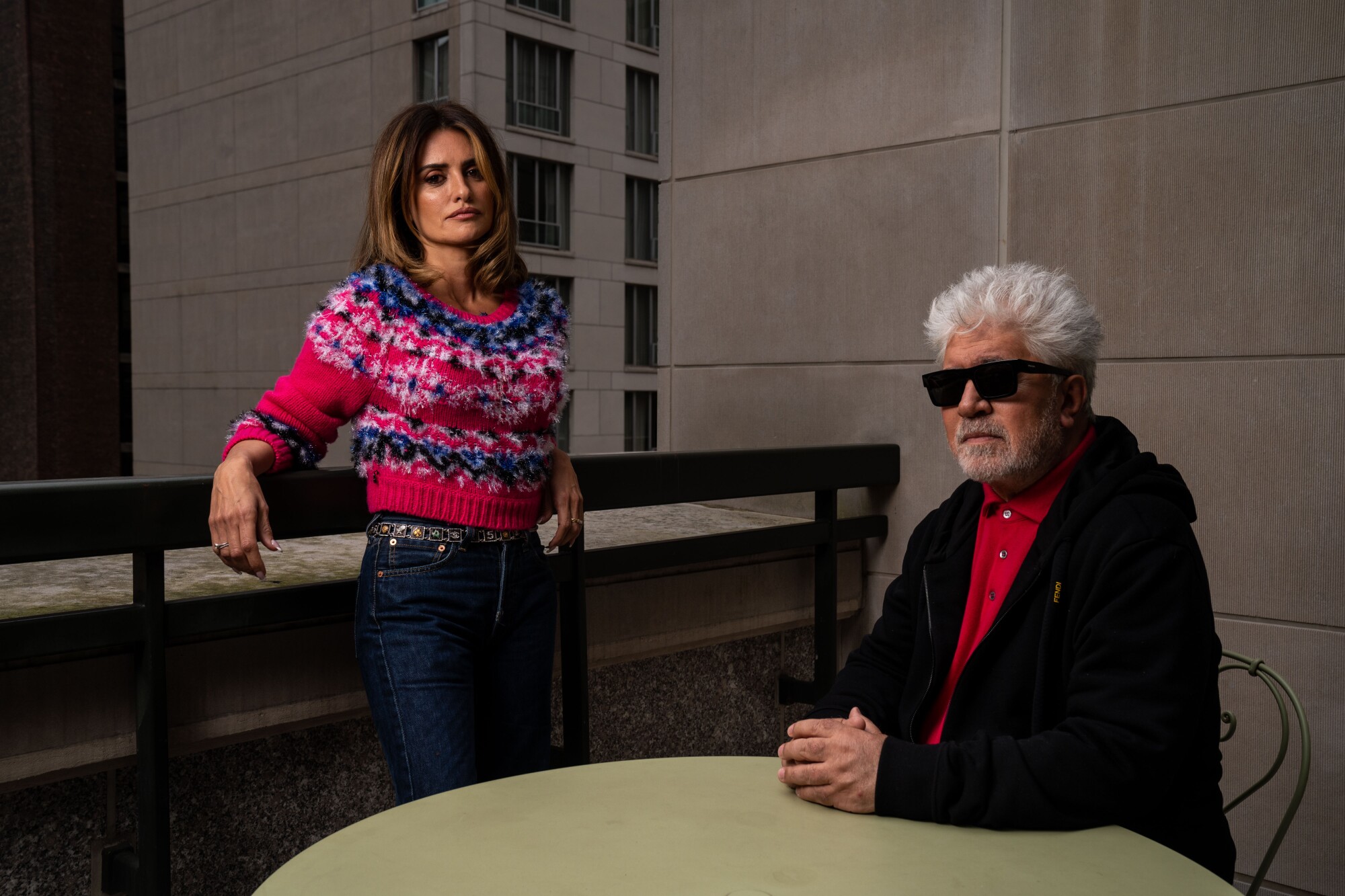 A woman in a red sweater leans against a railing while a man with white hair and sunglasses sits at a table outside 