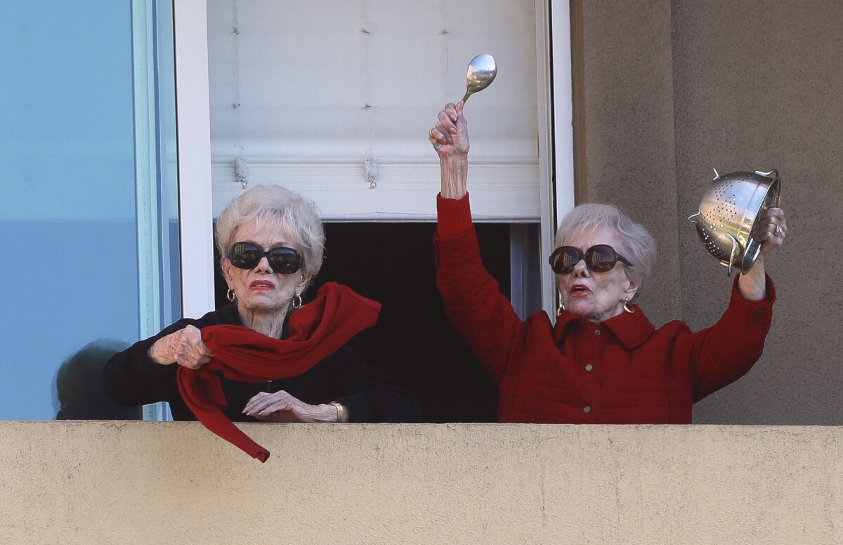 Joyce Kriesmer, 91, bangs a colander as she and her identical twin sister Jackie Voskamp waves a scarf on their balcony at the Vi at La Jolla Village senior living complex in La Jolla on Wednesday. Every weekday afternoon, the high-rise complex hosts a balcony pep rally for its residents who are sheltering in place because of the COVID-19 pandemic.